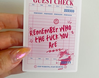 May include: A pink and white sticker resembling a guest check. The sticker features the words "GUEST CHECK" at the top, with lines for details. Bold, pink text reads "REMEMBER WHO THE FUCK YOU ARE." Includes a lipstick kiss mark and the phrase "STOP WORRYING, START BELIEVING."