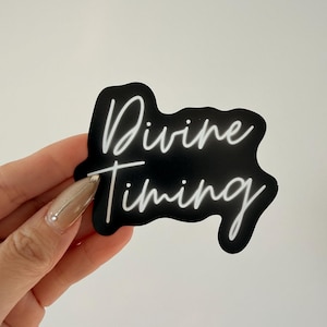 May include: A black sticker with the words "Divine Timing" in white cursive script. The sticker has a black border and is held against a white background. The sticker is a motivational or inspirational item.