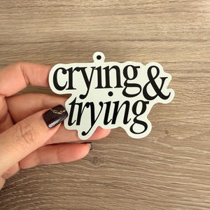 May include: A white sticker with the words "crying & trying" in a black, cursive font. The sticker is held by a hand against a wooden background. The sticker is a humorous, relatable statement.