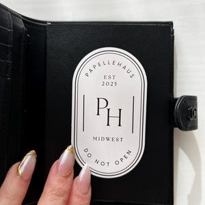 May include: A black leather wallet with a white sticker that says "Papellehaus Est 2025 PH Midwest Do Not Open".