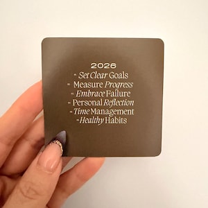 May include: A square, brown card with the year 2026 printed at the top. The card lists goals: Set Clear Goals, Measure Progress, Embrace Failure, Personal Reflection, Time Management, and Healthy Habits. The card is held by a person.
