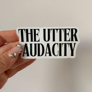 Może przedstawiać: Biała naklejka z napisem "THE UTTER AUDACITY" pogrubioną, czarną czcionką. Naklejka jest prostokątna z zaokrąglonymi rogami.