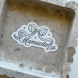 May include: A white sticker featuring a black bow and the word "Mamacita" in cursive writing. The sticker is placed on a grey, textured surface with a scalloped edge.