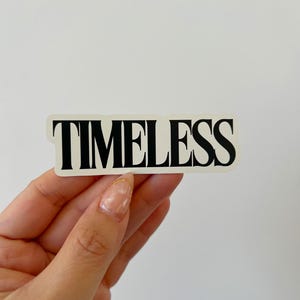 May include: A rectangular sticker with the word "TIMELESS" in bold, black capital letters. The sticker is held against a white background. The sticker's font is a classic serif style.