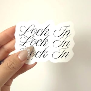 May include: A white sticker with the words "Lock In" printed three times in elegant black script. The sticker is held by a hand with manicured nails against a plain white background.