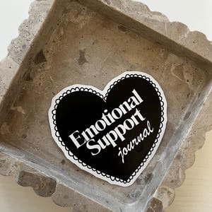 Op de afbeelding: Een zwarte sticker in de vorm van een hart met witte tekst die "Emotional Support Journal" luidt op een grijs stenen oppervlak.