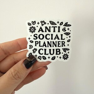 May include: A white sticker with the text "ANTI SOCIAL PLANNER CLUB" in a bold, black font. The sticker is surrounded by black floral and leaf designs. The sticker is held by a hand with a dark nail polish.