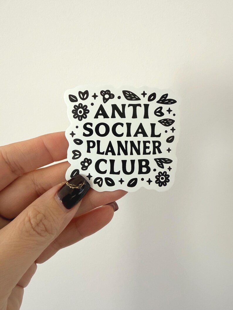 May include: A white sticker with the text "ANTI SOCIAL PLANNER CLUB" in a bold, black font. The sticker is surrounded by black floral and leaf designs. The sticker is held by a hand with a dark nail polish.