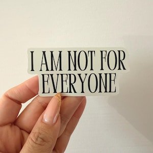May include: A rectangular sticker with the text "I AM NOT FOR EVERYONE" in black, sans-serif font. The sticker is held by a hand with a long, manicured nail.