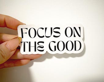 May include: A white sticker with the text "FOCUS ON THE GOOD" in black, stylized font. The sticker is rectangular with rounded corners. The sticker is being held up by a hand.