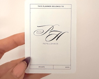 Pode incluir: Um cartão branco com o texto "THIS PLANNER BELONGS TO" no topo. Abaixo, as elegantes iniciais cursivas "PH" são exibidas, seguidas por "PAPELLEHAUS". A seção inferior diz "PRIVATE" e "20206".
