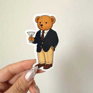 Pu&ograve; includere: Un adesivo con un orso dei cartoni animati in blazer blu navy, pantaloni color cachi e scarpe marroni, che tiene in mano un martini. L'orso indossa una cravatta a righe rosse e blu navy. L'adesivo &egrave; tenuto in mano da una mano con unghie lunghe e decorate.