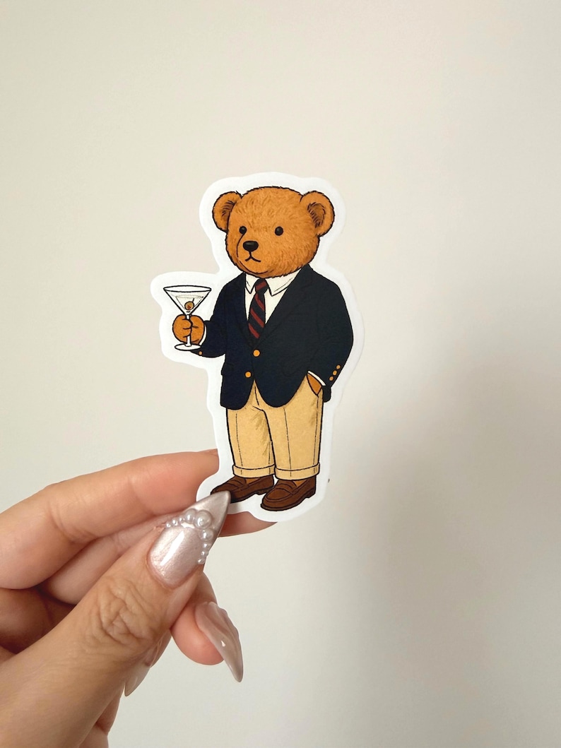 Pu&ograve; includere: Un adesivo con un orso dei cartoni animati in blazer blu navy, pantaloni color cachi e scarpe marroni, che tiene in mano un martini. L'orso indossa una cravatta a righe rosse e blu navy. L'adesivo &egrave; tenuto in mano da una mano con unghie lunghe e decorate.