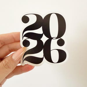 May include: A black and white sticker with the numbers "2026" in a bold, stylized font. The sticker is held against a white background. The numbers have a rounded, decorative design.