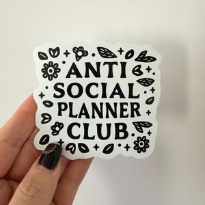 May include: A white sticker with black text that reads "ANTI SOCIAL PLANNER CLUB." The sticker is surrounded by black floral and leaf designs. The sticker is held by a hand with a dark manicure.