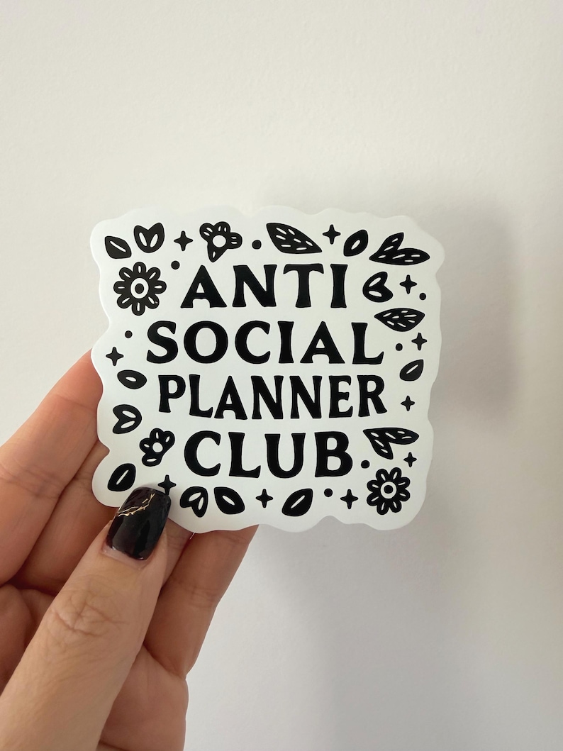 May include: A white sticker with black text that reads "ANTI SOCIAL PLANNER CLUB." The sticker is surrounded by black floral and leaf designs. The sticker is held by a hand with a dark manicure.