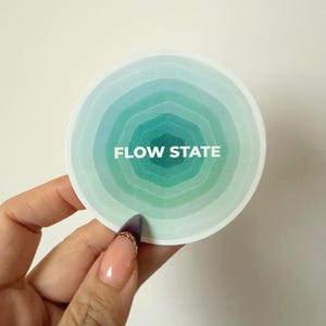 May include: A round sticker with a gradient design of blue and green concentric circles. The text "FLOW STATE" is centered in white. The sticker is held by a hand with a painted nail.