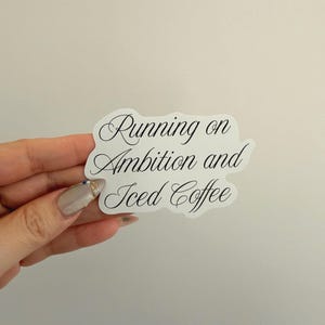 May include: A white sticker with the text "Running on Ambition and Iced Coffee" in elegant black script. The sticker is held against a plain, off-white background.