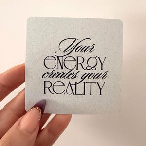 May include: A square, light gray sticker with the text "Your energy creates your reality" in black script. The sticker is held by a hand with a painted fingernail.