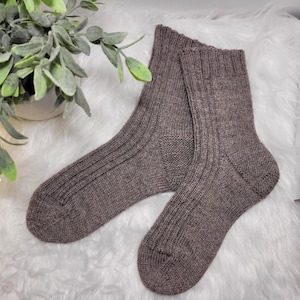 May include: A pair of brown ribbed knit socks. The socks are made of a soft, textured yarn and have a classic design. The socks are displayed on a white, fluffy surface with a green plant in the background.