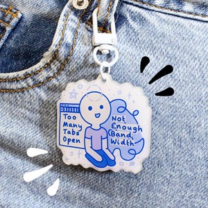 Peut inclure: Un porte-clés en acrylique transparent avec un fermoir en métal blanc, accroché à un jean bleu. Le porte-clés présente une illustration de dessin animé bleu et blanc avec le texte "Too Many Tabs Open" et "Not Enough Band Width."