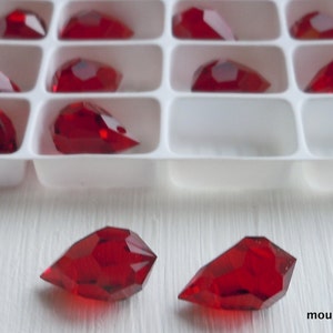 May include: Red faceted teardrop-shaped beads, some in a white plastic tray, others loose. The beads have a glossy finish and are arranged on a white surface. The text "mountainshadowbeads" is visible.
