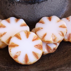 May include: A collection of white and orange flower-shaped beads. Each bead has a white base with orange starburst patterns. The beads are arranged on a dark, textured surface, creating a visually appealing display for jewelry making or crafting.