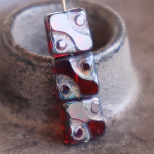 May include: A stack of three rectangular glass beads with a red and white design. Each bead has a unique pattern with a circular accent. The beads are strung together on a thin gold wire, perfect for jewelry making.