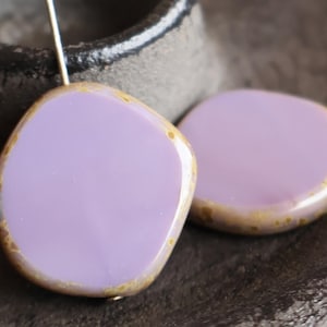 May include: Two lavender-colored, irregular-shaped beads with a gold-colored edge. The beads are smooth and shiny, with a slightly rounded shape. The beads are likely for jewelry making.