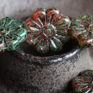 May include: Close-up of several flower-shaped beads. The beads have a textured surface with a mix of green, red, and brown colors. The beads are arranged in a row, with some of them partially visible. The beads are placed on a dark, textured surface.