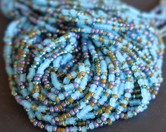 May include: A close-up of a collection of beaded strands. The beads are primarily light blue, with accents of purple, gold, and silver. The beads are small and round, creating a textured appearance. The strands are bunched together, suggesting a necklace or other jewelry.