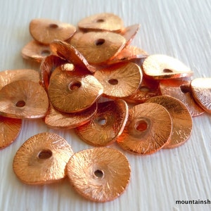May include: A pile of copper-colored metal beads with a hole in the center of each bead. The beads have a textured surface and are arranged in a random pattern.
