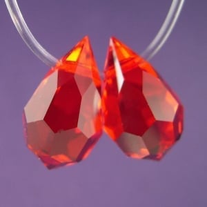 以下が含まれることがあります： 透明な糸に吊るされた、ファセット加工された赤色のしずく型のクリスタルビーズ2つ。クリスタルは、複数のファセットで光を捉え、宝石のような輝きを放っています。背景はソフトパープルです。
