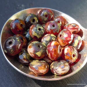 May include: A selection of artisan beads presented in a silver spoon. The beads display a blend of colours, such as red, brown, gold, and white. Each bead is pierced with a hole for stringing. The beads have a glossy finish, possibly made of glass or a similar material.