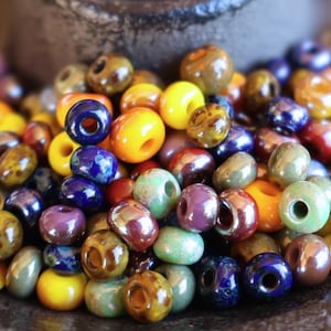 May include: A collection of colorful beads in various sizes and shades, including yellow, blue, green, and brown. The beads have a glossy finish and are arranged in a pile, suggesting they are for jewelry making or crafts.
