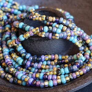 May include: Close-up of colorful beaded necklaces in shades of blue, yellow, purple, and black. The beads are small and round, strung together to create a layered effect. The necklaces are resting on a dark, textured surface.