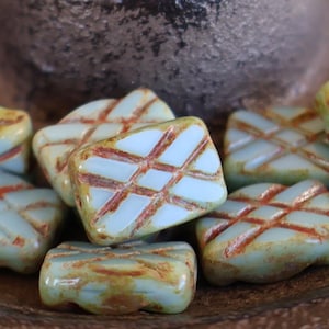 May include: A collection of square-shaped beads with a light blue base and a crisscross pattern in a reddish-brown hue. The beads have a slightly rounded shape and a glossy finish, suggesting they are made of glass or a similar material. They are arranged on a dark brown surface.