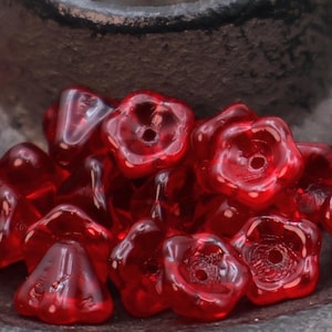 May include: Close-up of a collection of translucent red glass beads. Some beads are flower-shaped with a central hole, while others are triangular. The beads are scattered on a dark, textured surface.