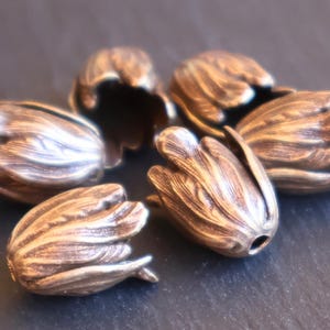 May include: A collection of six copper-toned metal bead caps shaped like closed flower buds. Each bead cap features a textured, ribbed surface with a dark patina finish, highlighting the intricate petal-like details and a central hole for stringing.