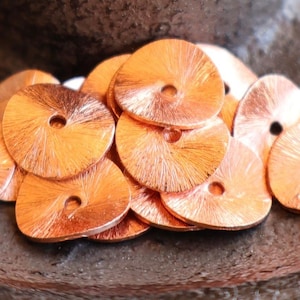 May include: A collection of copper-colored, circular metal sequins with a central hole. The sequins have a textured, radial pattern emanating from the center. They are arranged on a dark, textured surface, creating a warm, metallic aesthetic. Ideal for craft projects.