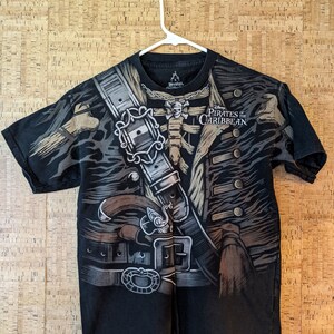 May include: Black t-shirt featuring a detailed graphic of a pirate's vest and jacket, complete with buckles, a pistol, and a skull and crossbones. The words "Disney Pirates of the Caribbean" are printed on the right side of the shirt.