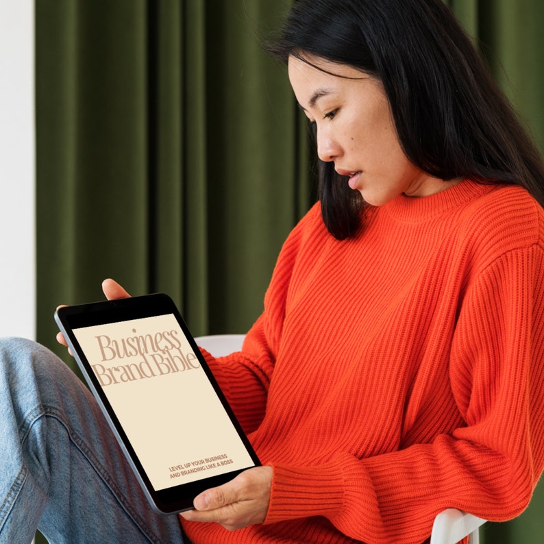 May include: A person in a red sweater is reading a digital tablet. The tablet screen displays the text "Business Brand Bible" and "Level up your business and branding like a boss."