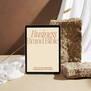 May include: A digital tablet displaying a mock-up of a "Business Brand Bible" guide with the text "Level Up Your Business and Branding Like a Boss". The tablet is on a light brown surface with a white fabric and a brown rock in the background.