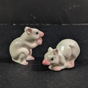 May include: Two white ceramic mouse figurines. One mouse is sitting up with its paws together, while the other mouse is lying down with its head turned to the side. Both mice have pink noses and ears.