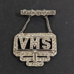 May include: A silver-toned pin with a chain and a rectangular pendant. The pendant is decorated with rhinestones and has the letters "VMS" in the center.