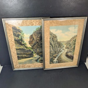 May include: Two rectangular framed prints featuring landscape scenes of rocky canyons with winding roads and a river. Each print is encased in a thin silver metal frame with a wide, textured tan matting surrounding the detailed nature imagery.
