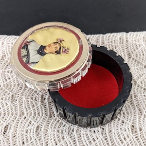 May include: An open, vintage black box with a clear, ridged lid. The lid features a portrait on a yellow background, framed by red. The interior is lined with red velvet. A decorative storage item.
