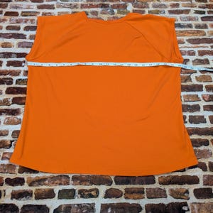 May include: A bright orange sleeveless top is displayed flat on a brick surface. A measuring tape is stretched across the garment, showing its width. The top's colour is a solid, vibrant orange.