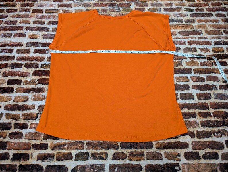 May include: A bright orange sleeveless top is displayed flat on a brick surface. A measuring tape is stretched across the garment, showing its width. The top's colour is a solid, vibrant orange.