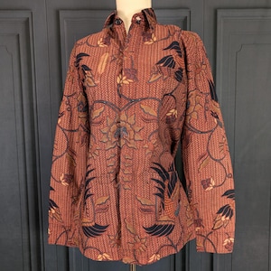 Peut inclure: Chemise à manches longues avec un motif batik. La chemise présente un fond brun rougeâtre avec un motif floral et de feuilles détaillé dans des tons de marron, bleu marine et beige. La chemise est présentée sur un mannequin.